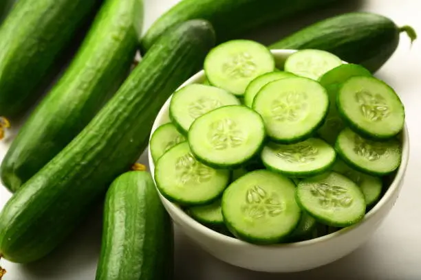 sliced cucumbers in a ceramic bowl with full cucumbers on a white background.
